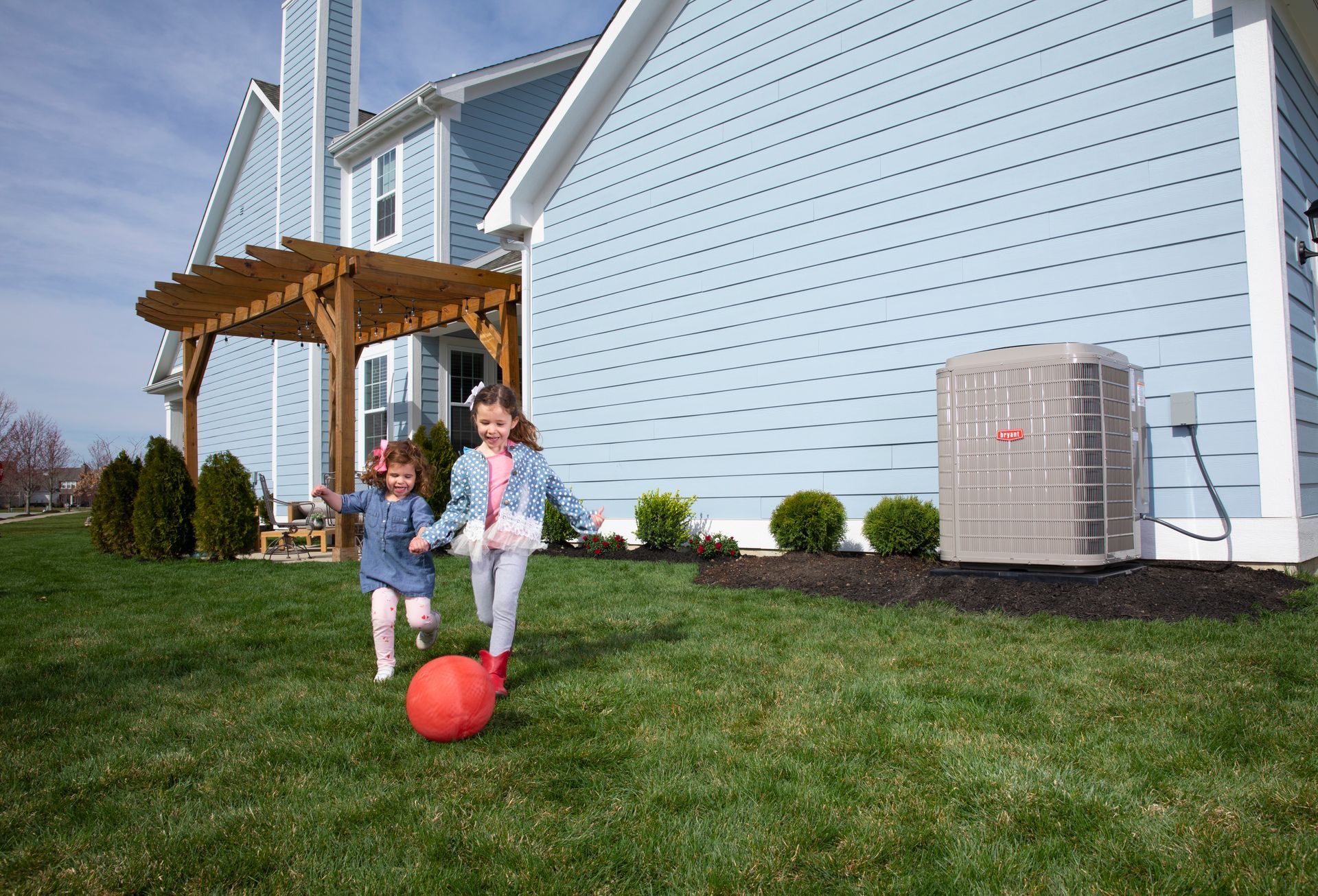 Two children run on a lawn, toward a blue house with an air conditioning unit.