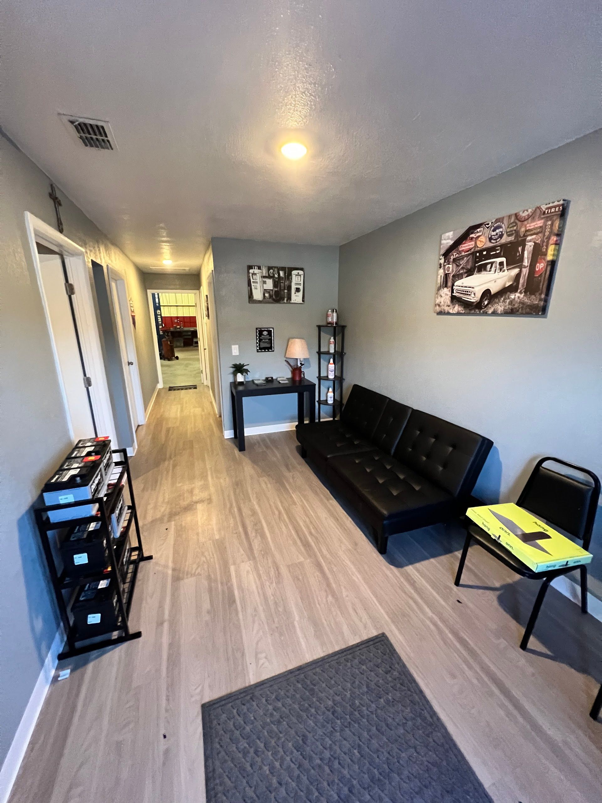 A hallway with light wood floors leading to another room, featuring a black sofa, a small table, and a chair. | Keep It Rollin’ Auto Repair LLC