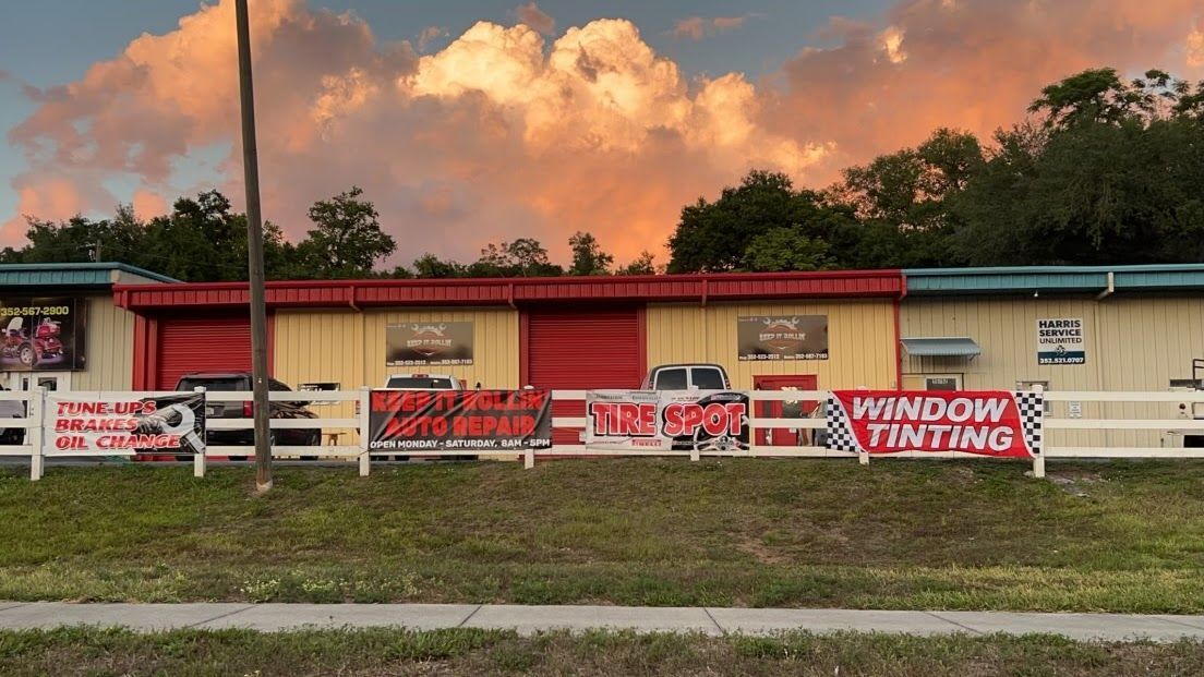 A tan commercial building with red bay doors and banners for auto services behind a white fence under a sunset sky. | Keep It Rollin’ Auto Repair LLC