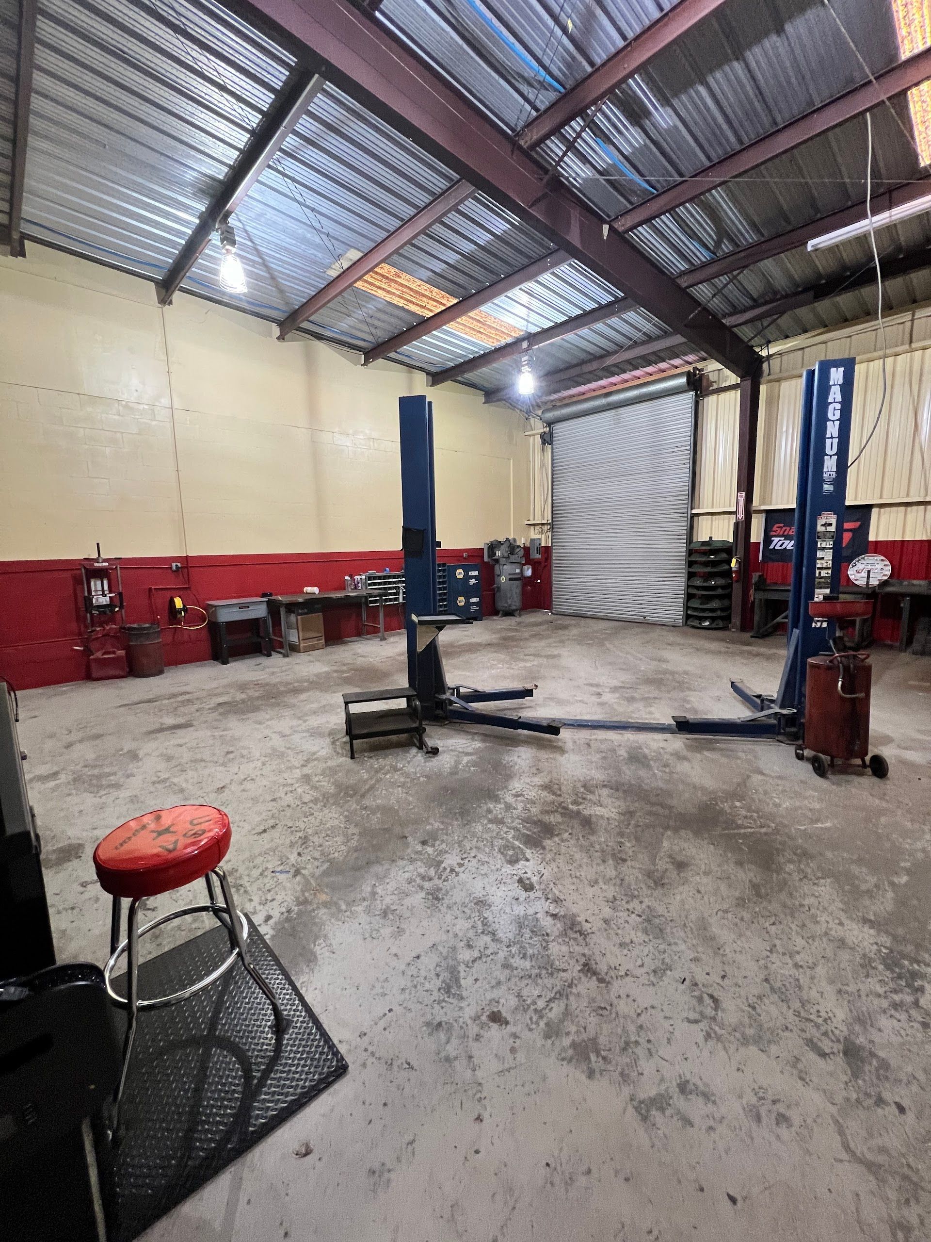 An empty auto repair shop with a vehicle lift in the center, a red stool in the foreground, and a roll-up garage door. | Keep It Rollin’ Auto Repair LLC