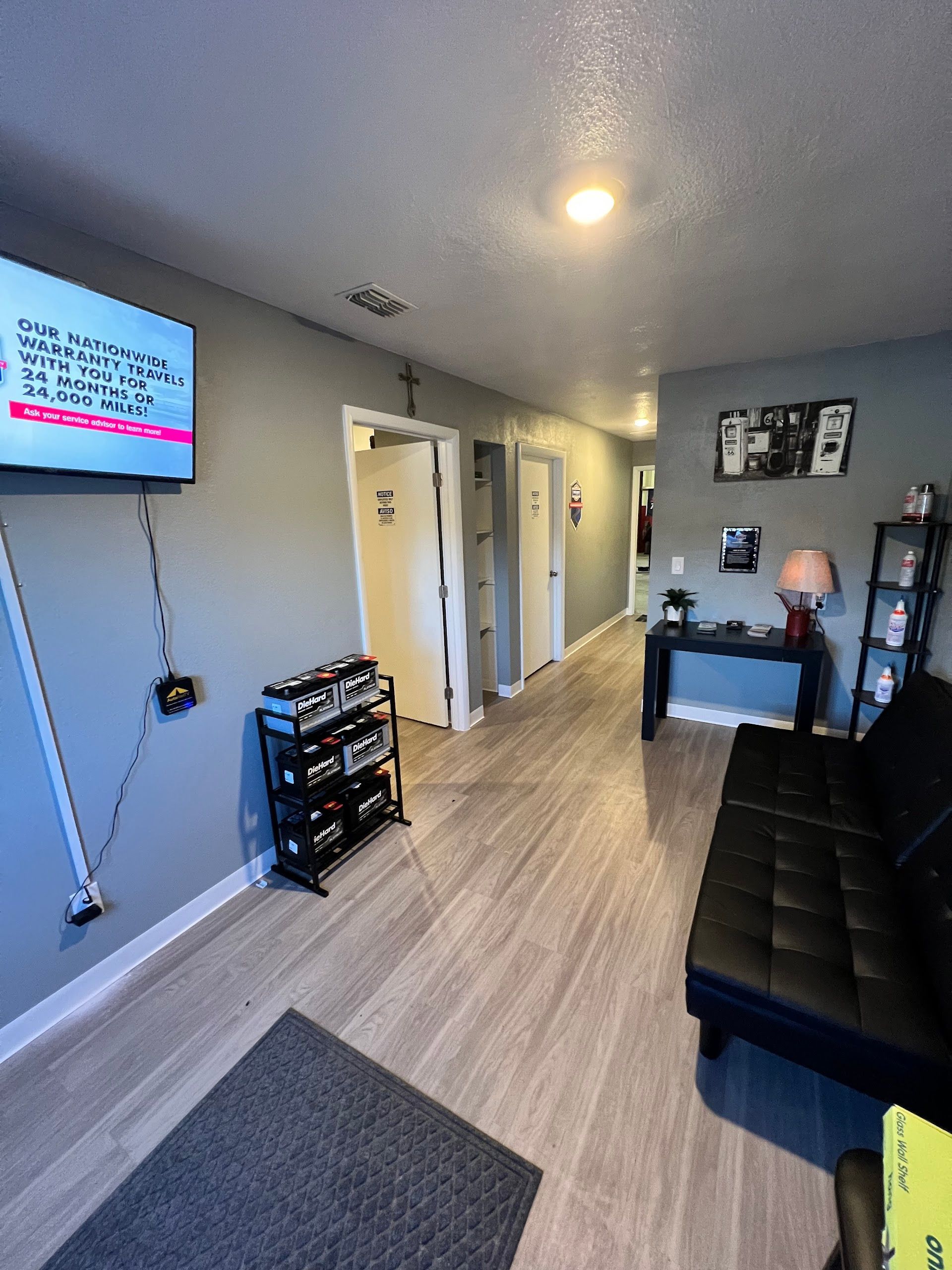 A room with grey walls, light wood flooring, a television on the wall, a shoe rack, a black sofa, and an entry hallway. | Keep It Rollin’ Auto Repair LLC