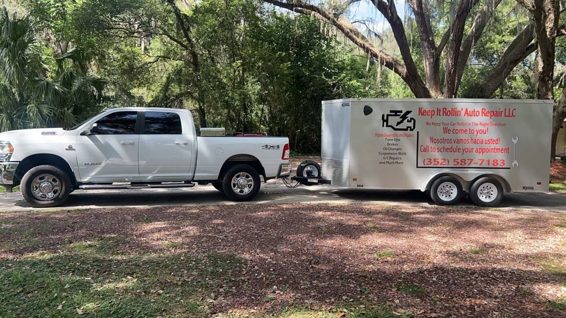 A white pickup truck parked in a wooded area, towing a silver, branded enclosed trailer with contact information. | Keep It Rollin’ Auto Repair LLC