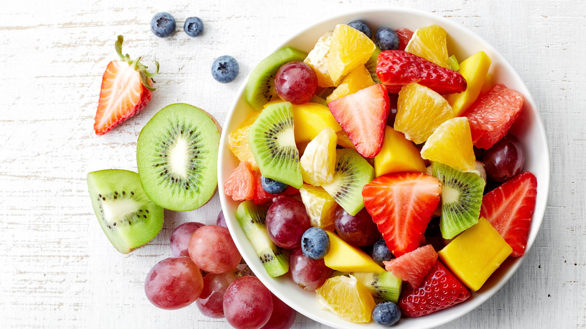 A bowl of fruit salad with kiwi , strawberries , grapes and blueberries on a table.