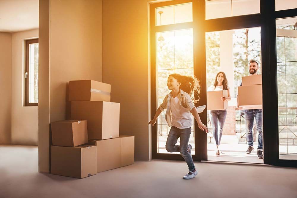 Family moving into a new home. A girl runs ahead, while parents carry boxes through the doorway. Sunlit interior.