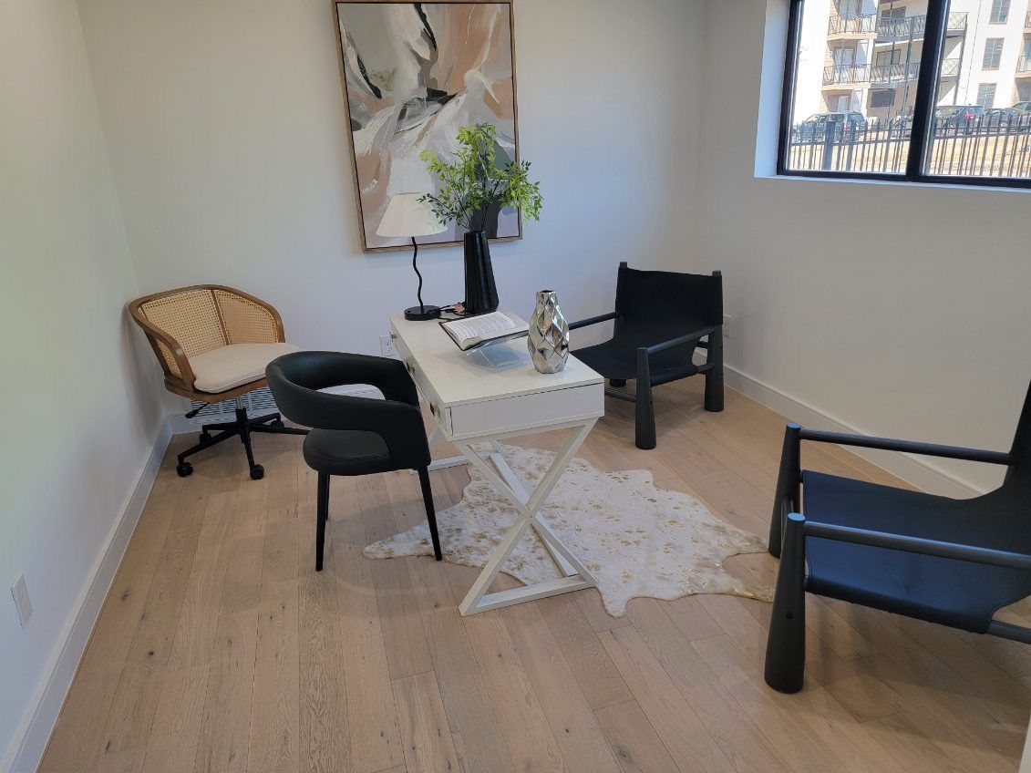 Office space with a white desk, three chairs, and abstract art. Light wood floors and a window are visible.