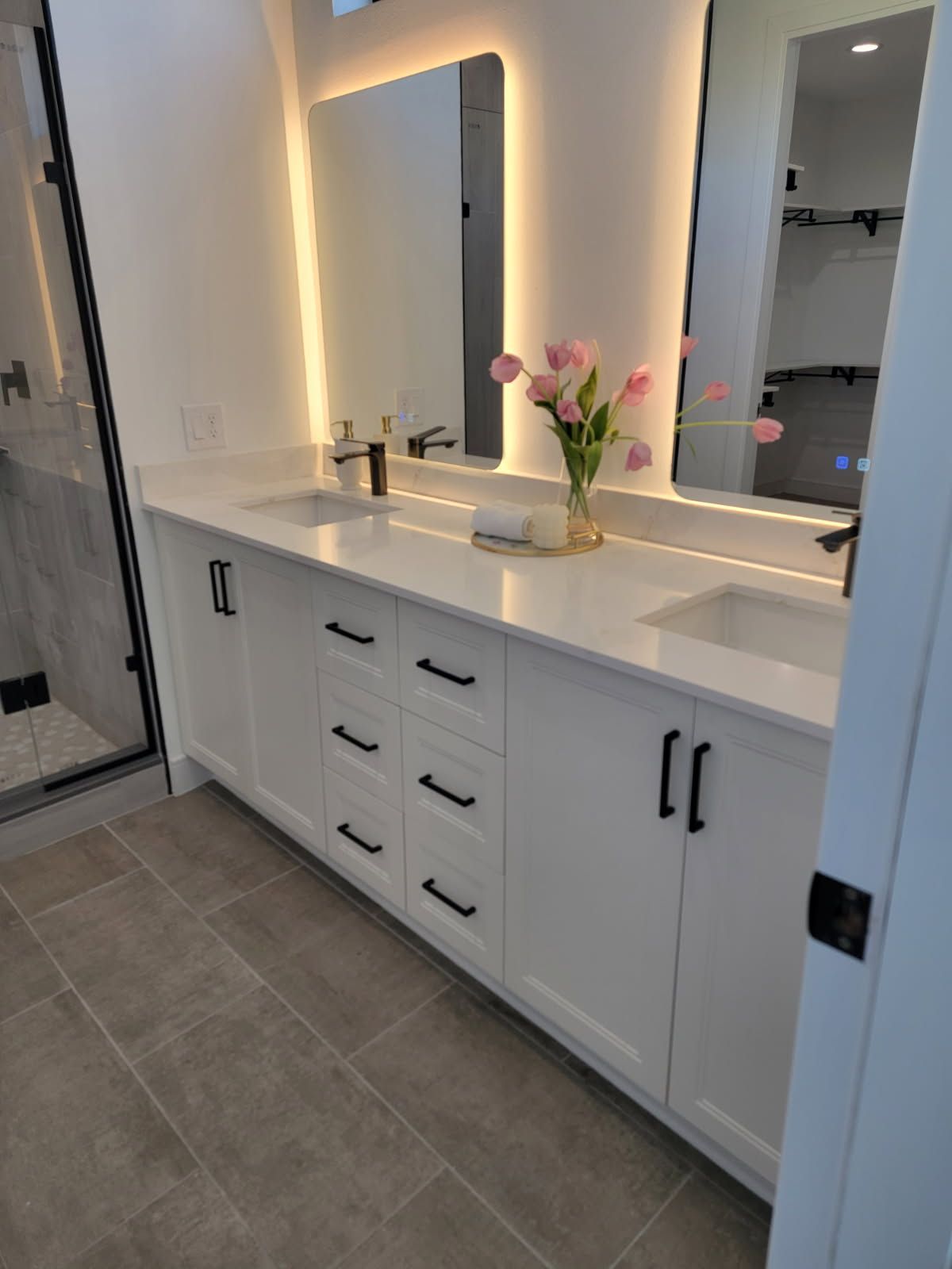White bathroom vanity with black hardware, two sinks, mirrors with backlighting, and a vase of pink tulips.