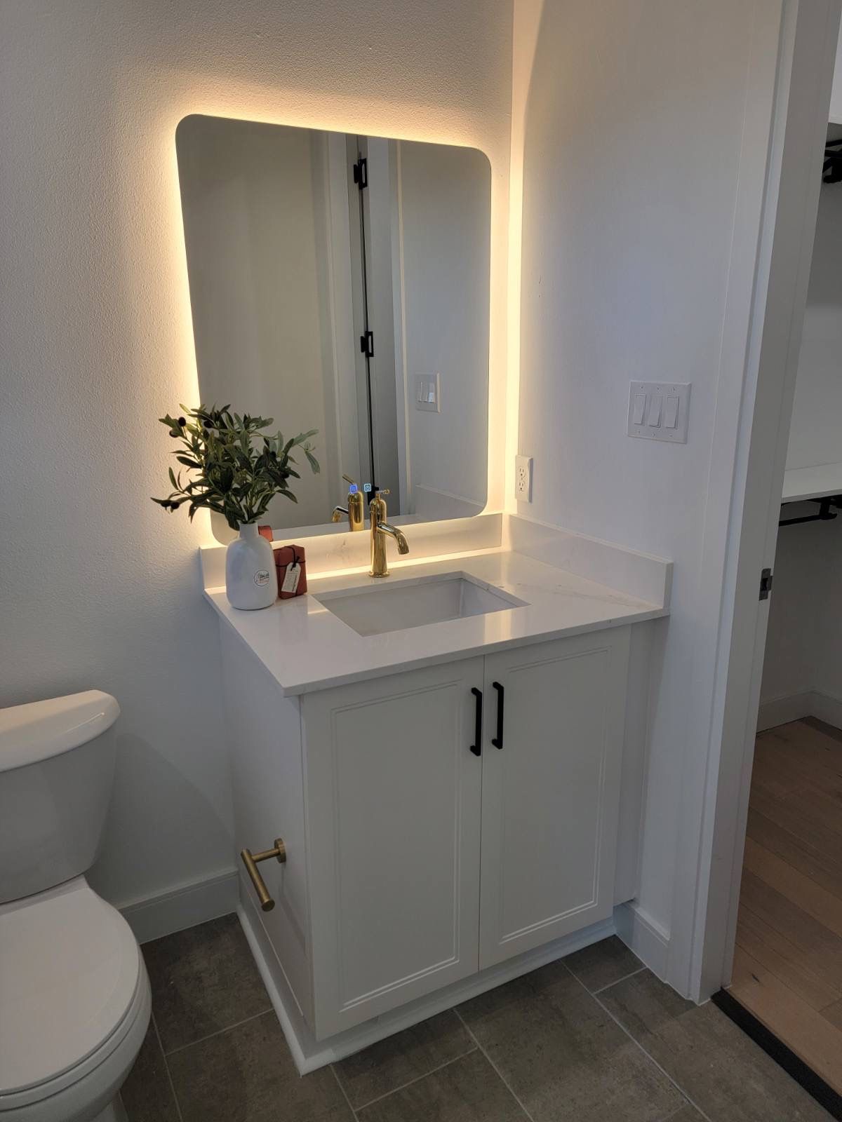 White bathroom vanity with lighted mirror, gold faucet and hardware, next to toilet.