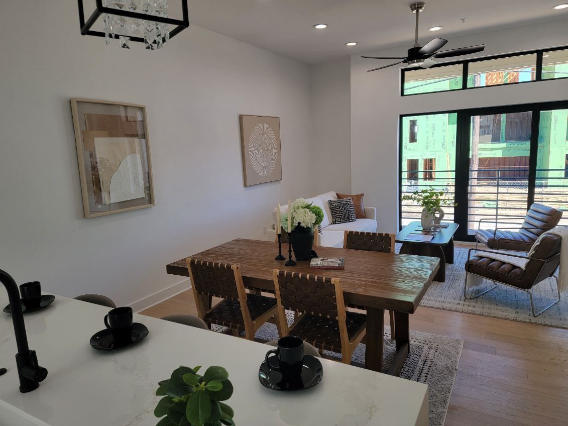 Dining area with wooden table, chairs, and art. Living area with sofa, chair, and large windows.
