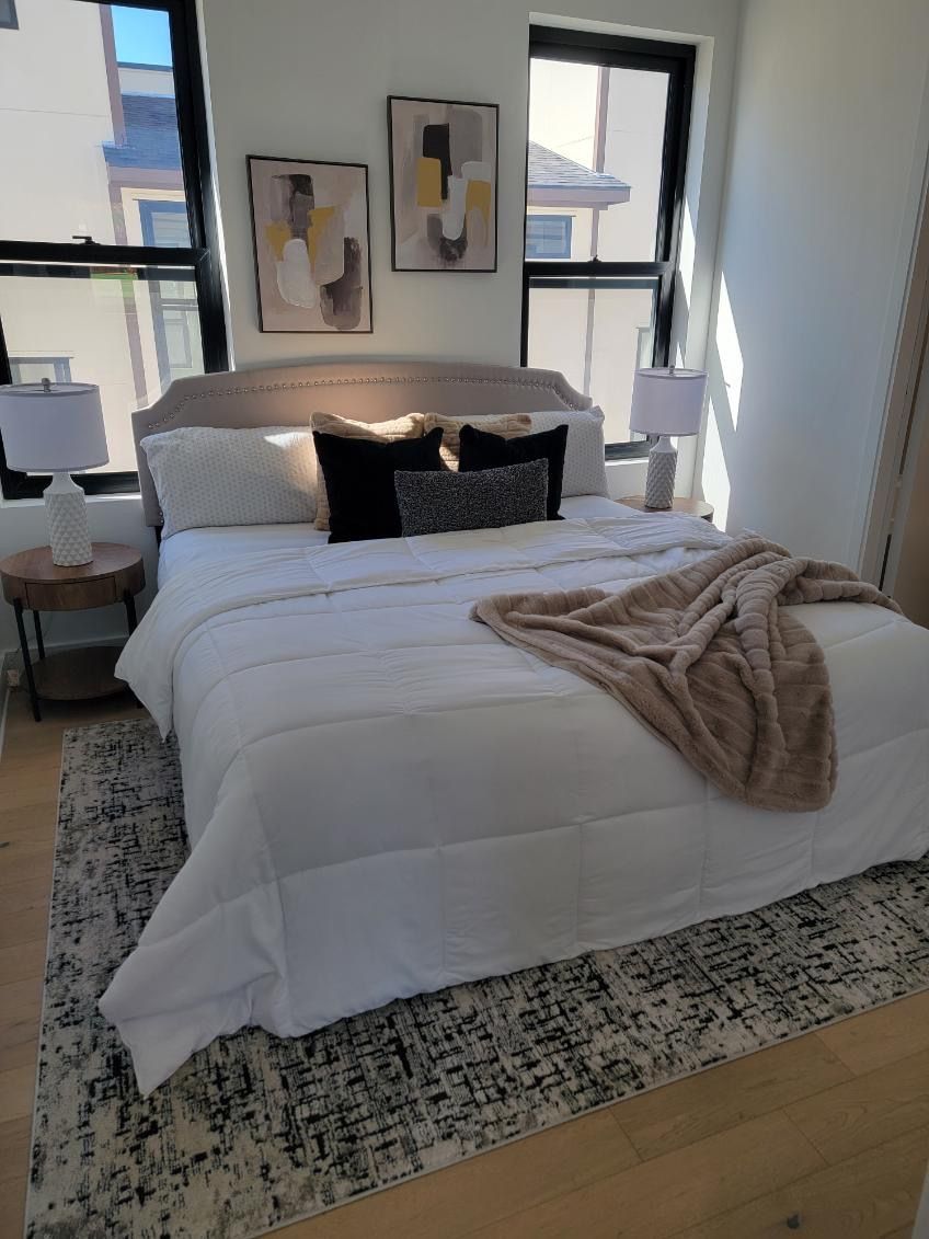 Bedroom with bed, pillows, rug, lamps, and artwork. Soft colors; sunlight streams in.
