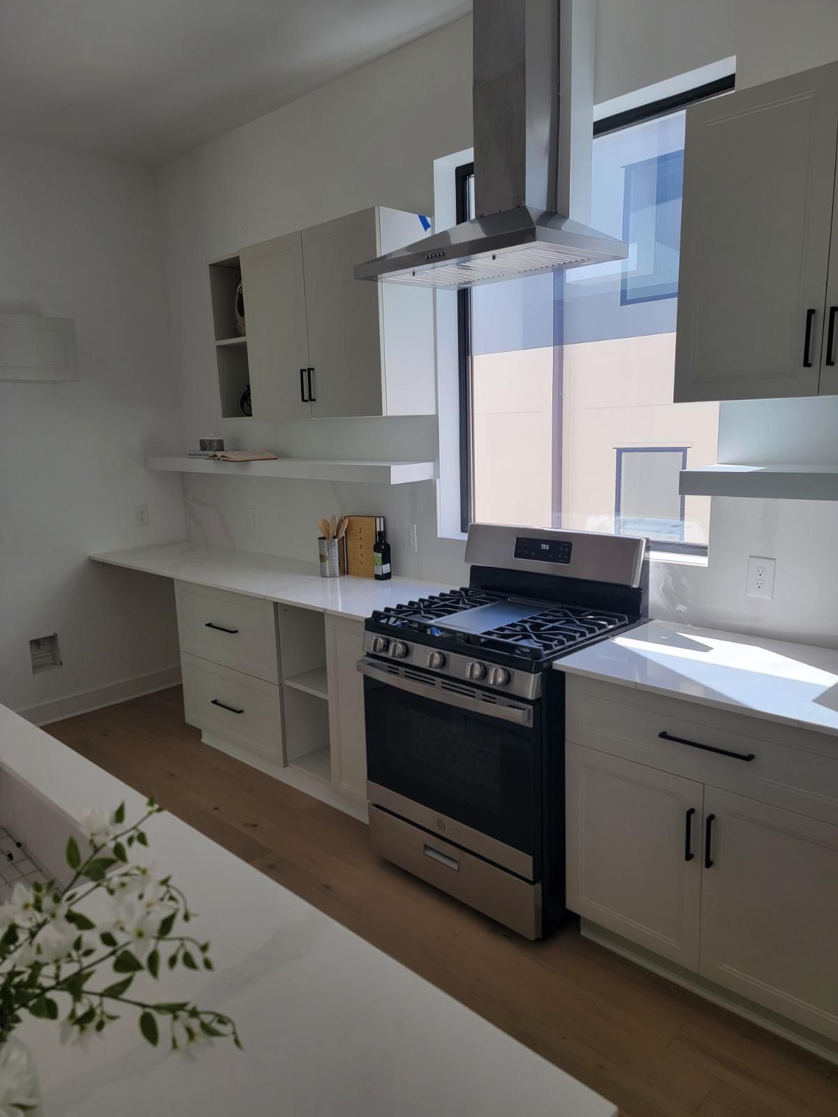 Modern white kitchen with stainless steel appliances and open shelving. Natural light streams from window.