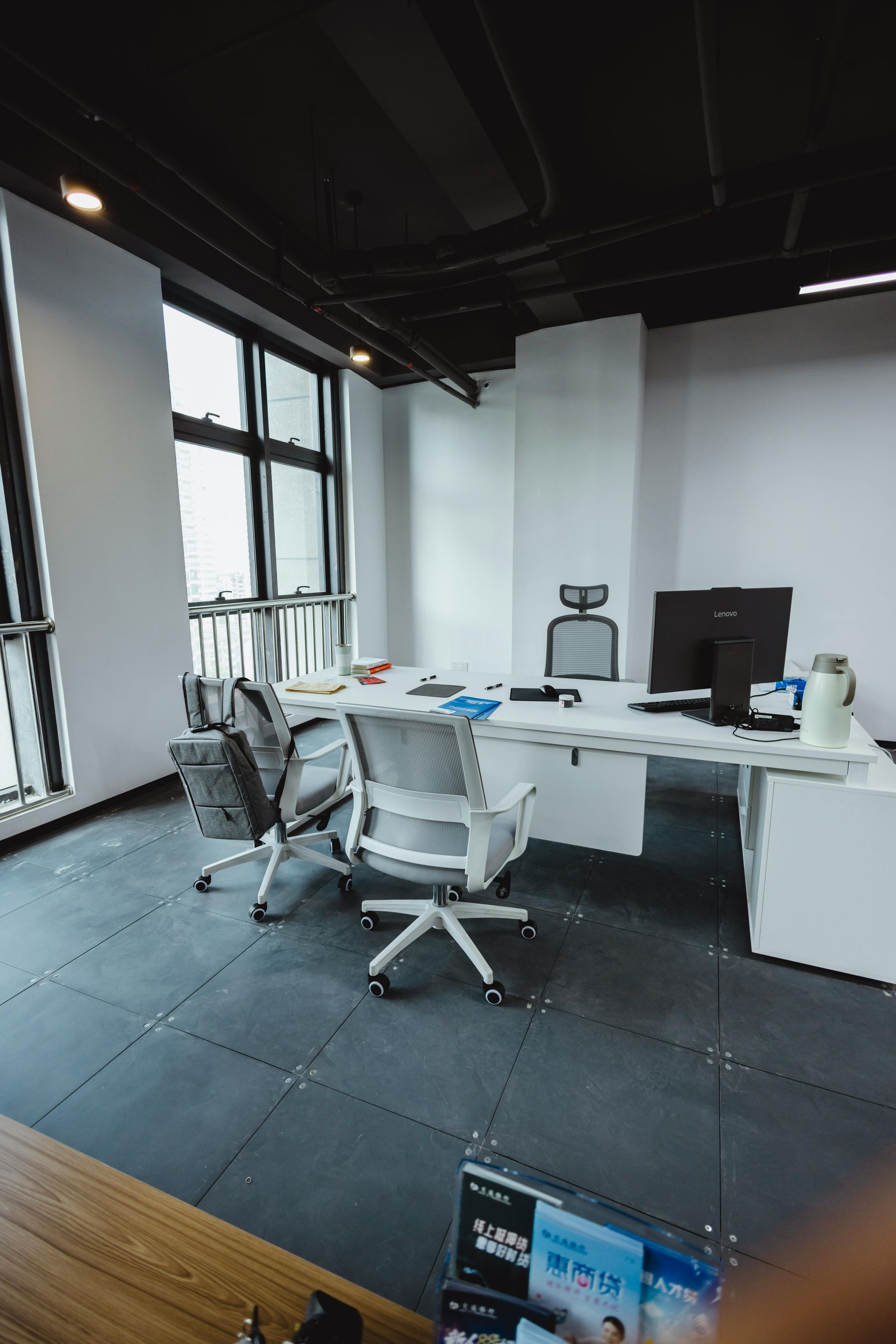 Office workspace with white desk, computer, and rolling chairs. Dark ceiling and floor with window and natural light.