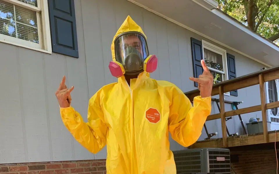 Person in yellow hazmat suit, respirator, flashing rock on fingers, standing in front of a house.