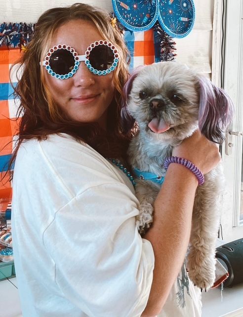 Woman in round blue sunglasses holding a small gray-and-white dog indoors.