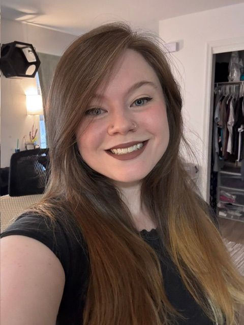 Smiling person taking a selfie in a bedroom, wearing a black shirt with long brown hair.