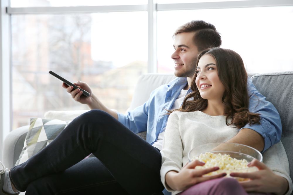 A Man And A Woman Are Sitting On A Couch Eating Popcorn And Watching Tv — Craig's TV/Antenna Services In Merewether, NSW