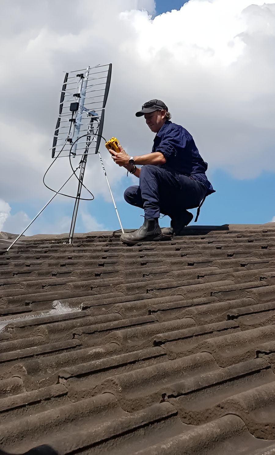 A Man Is Kneeling On Top Of A Roof Fixing An Antenna — Craig's TV/Antenna Services In Cameron Park, NSW