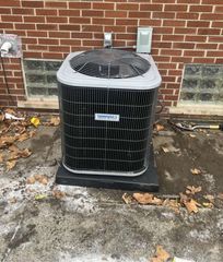 A residential HVAC condenser unit sits on a black pad against a brick wall with glass block windows.
