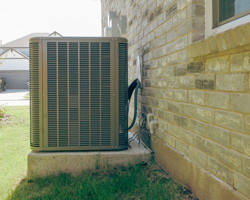 A tan residential air conditioning condenser unit sits on a concrete slab next to a light brick house wall.