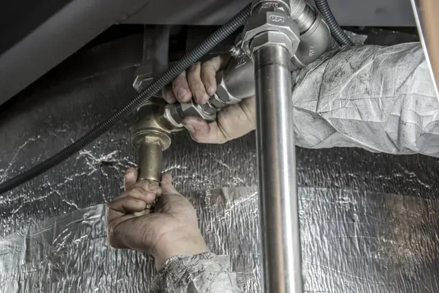 Les mains d'un plombier connectent des tuyaux métalliques dans un sous-sol, les tuyaux sont argentés et dorés, les murs sont isolés.