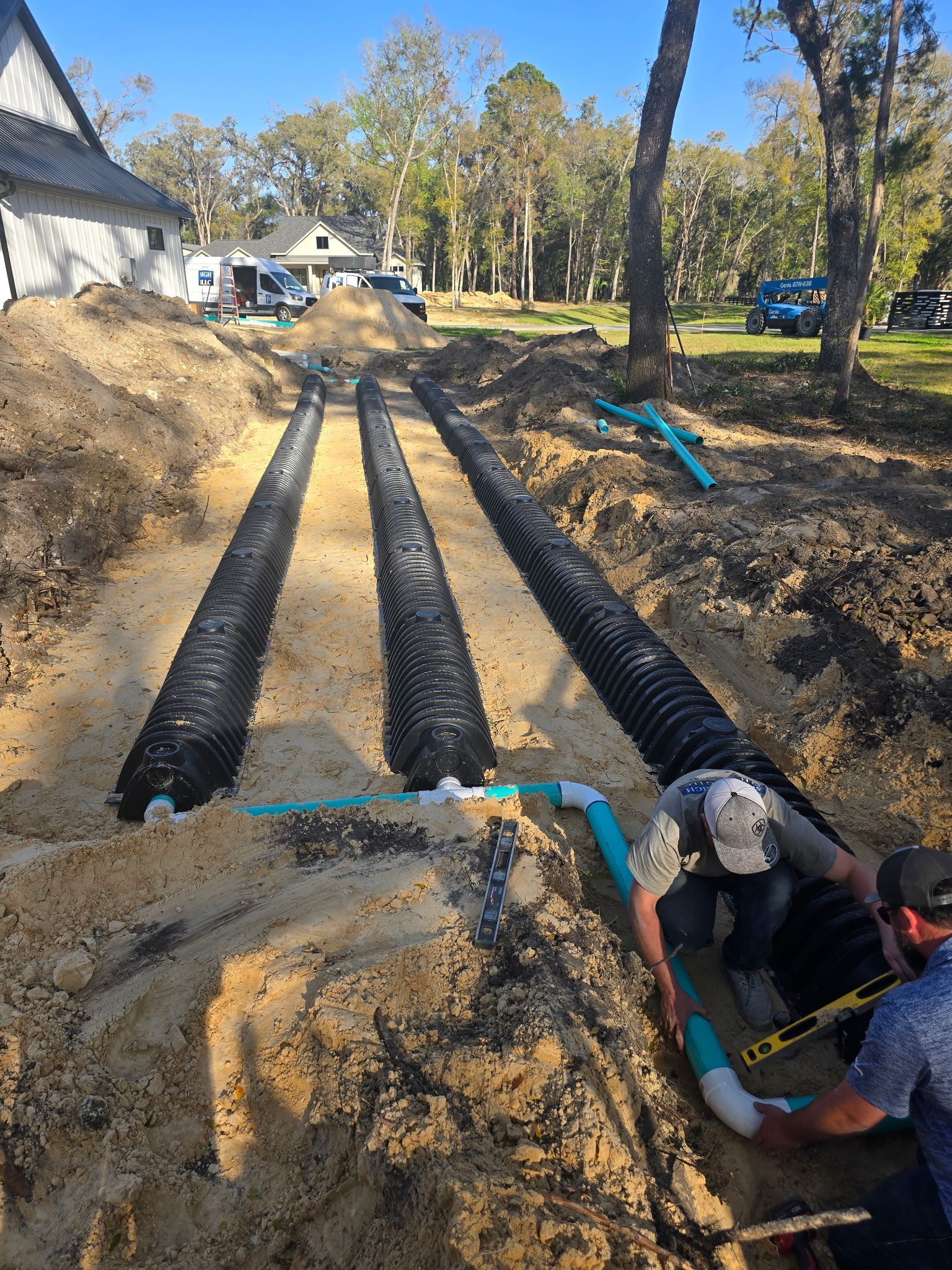 Workers installing drainage pipes in a dirt trench outdoors near a house, trees, and vehicles.