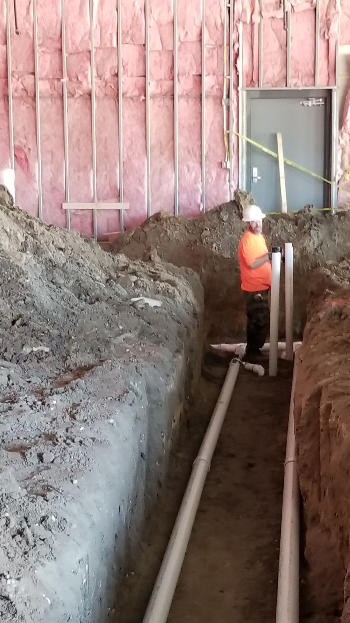 Construction worker installing pipes in a trench near a building with exposed pink insulation.