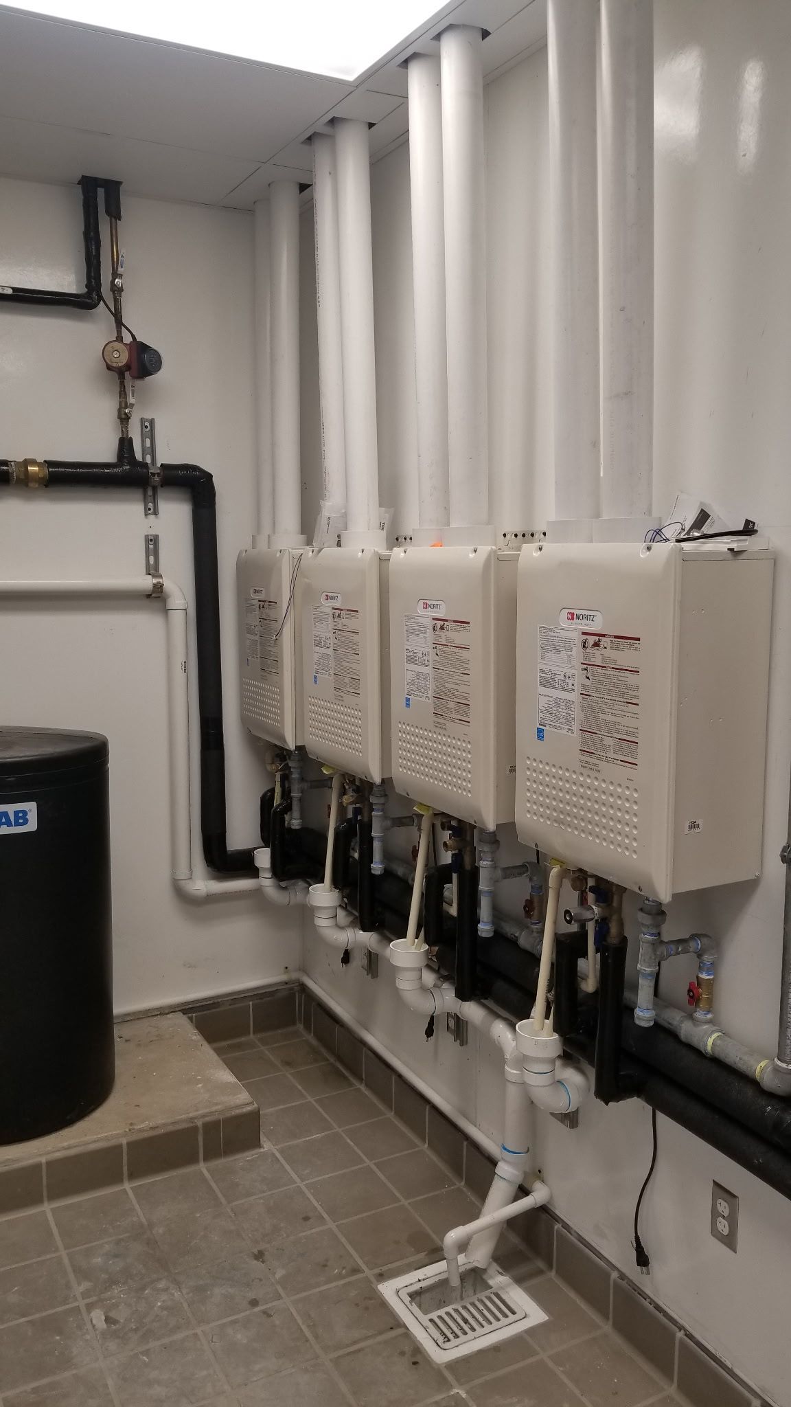 White water heaters, pipes, and a black tank in a utility room.