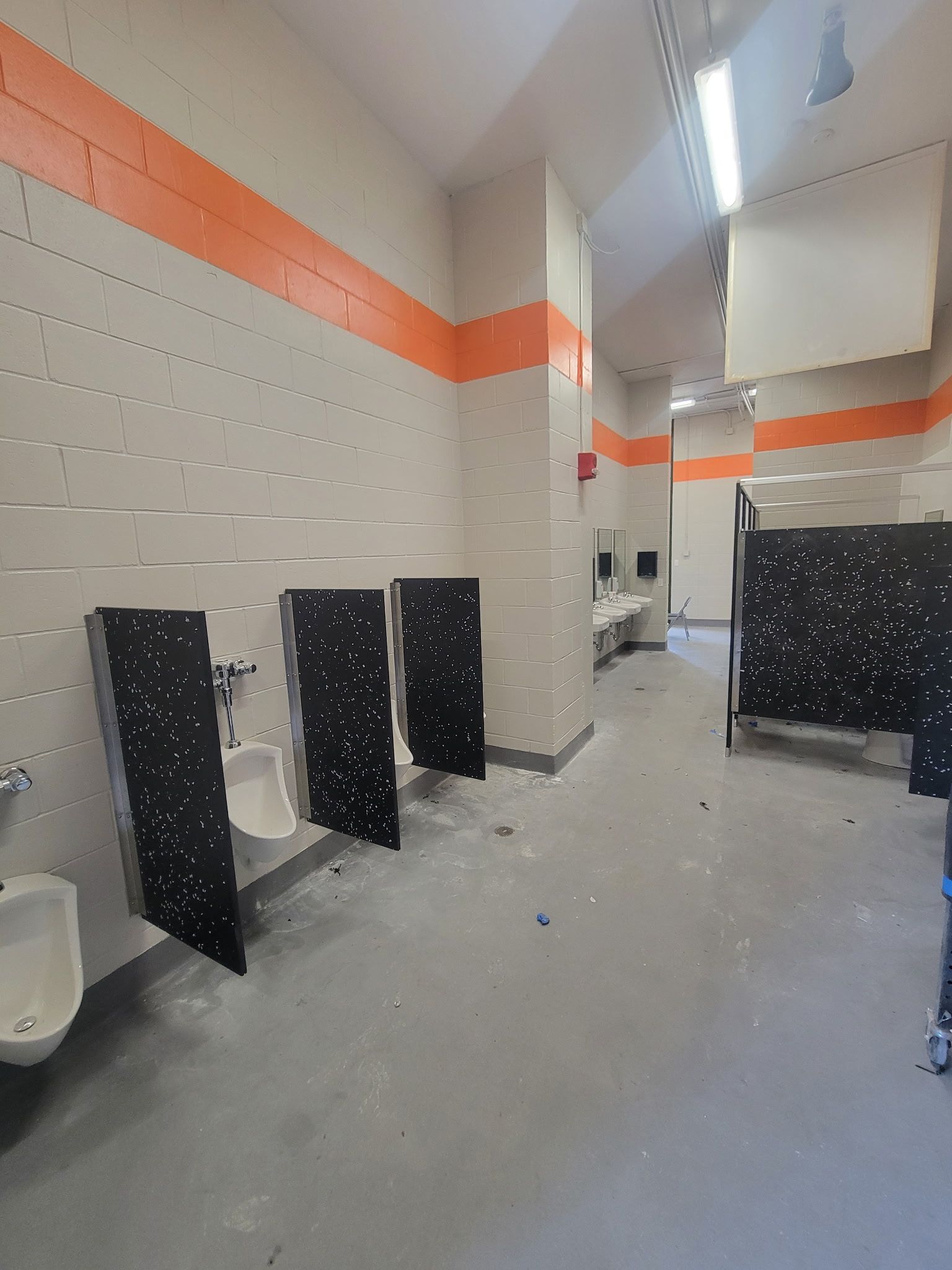 A public restroom with light gray walls, orange stripes, and black privacy partitions separating urinals and stalls.