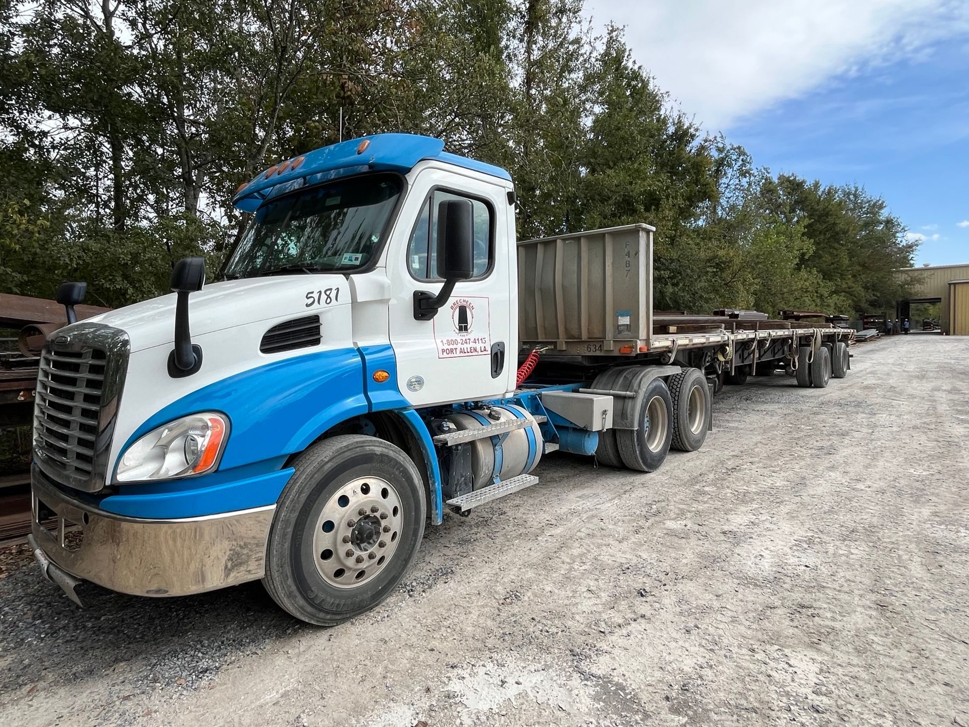 A blue and white semi truck with a flatbed trailer is parked in a gravel lot.
