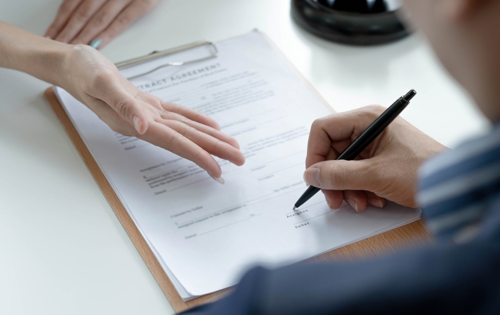Person signing a document on a clipboard, with another person's hand pointing at the document on a white table.