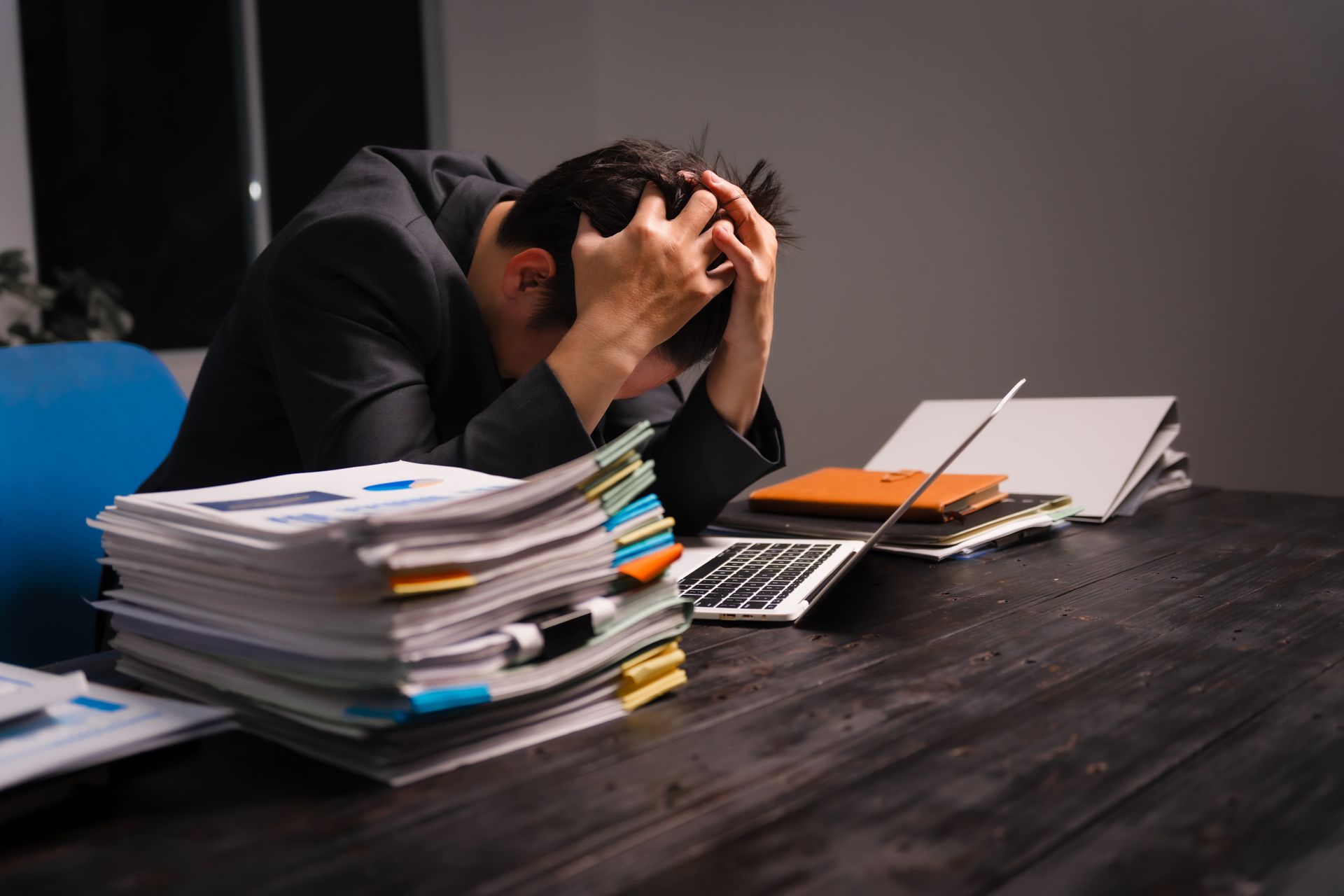 Man stressed at desk with stack of papers and laptop, hands on head.