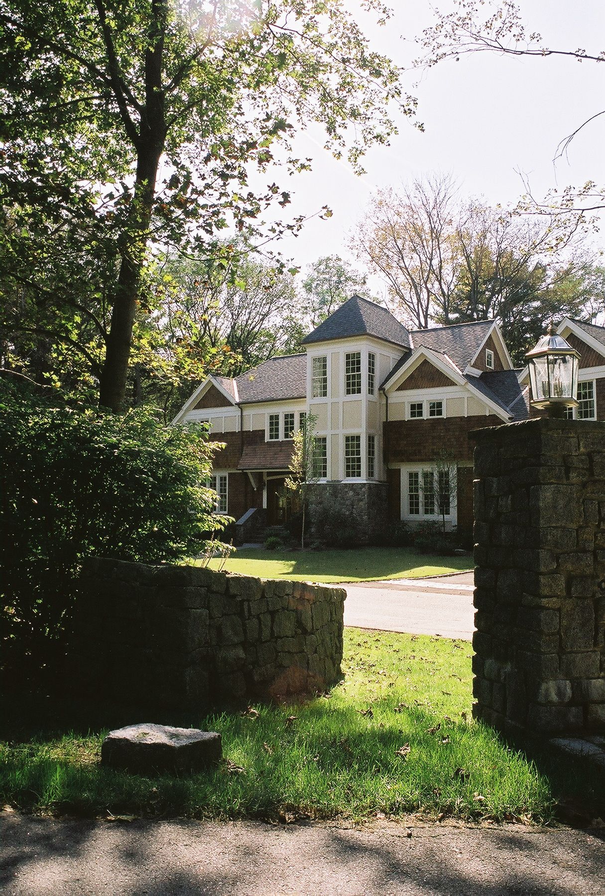 A two-story house with stone and light-colored siding, a turret, and a lawn behind a stone wall.
