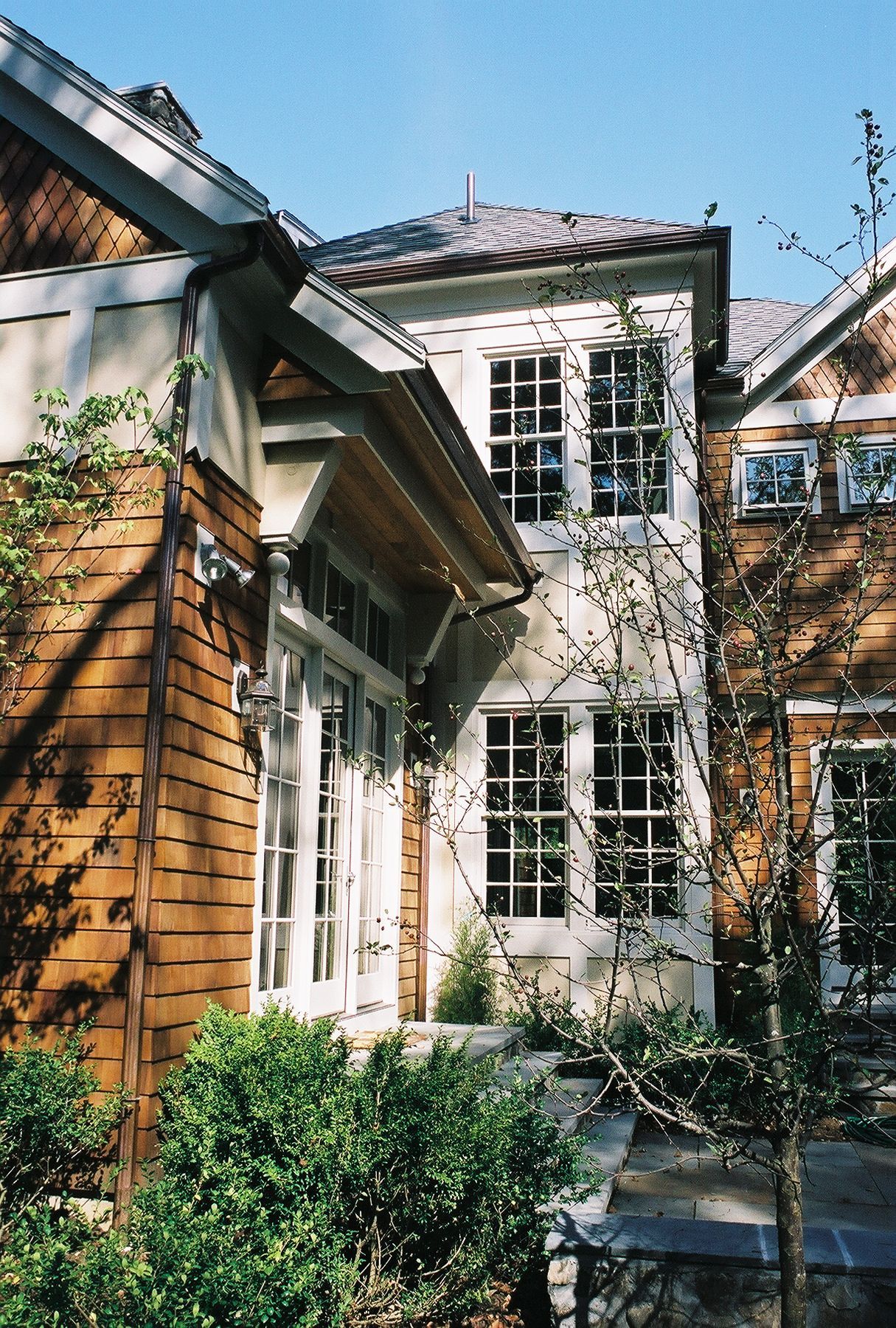 Brown-shingled house with white-framed windows, surrounded by greenery. Sunny day.