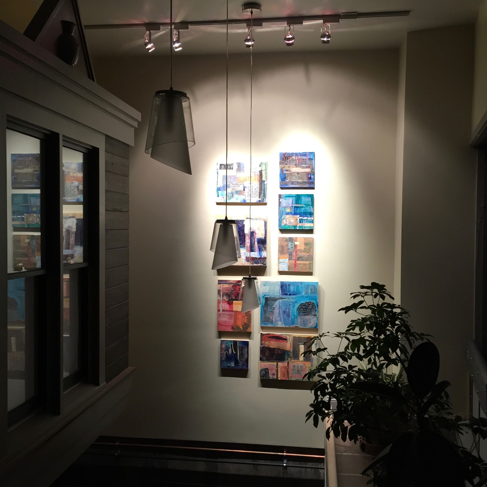 A wall with paintings illuminated by track lighting; a window on the left and a plant on the right.