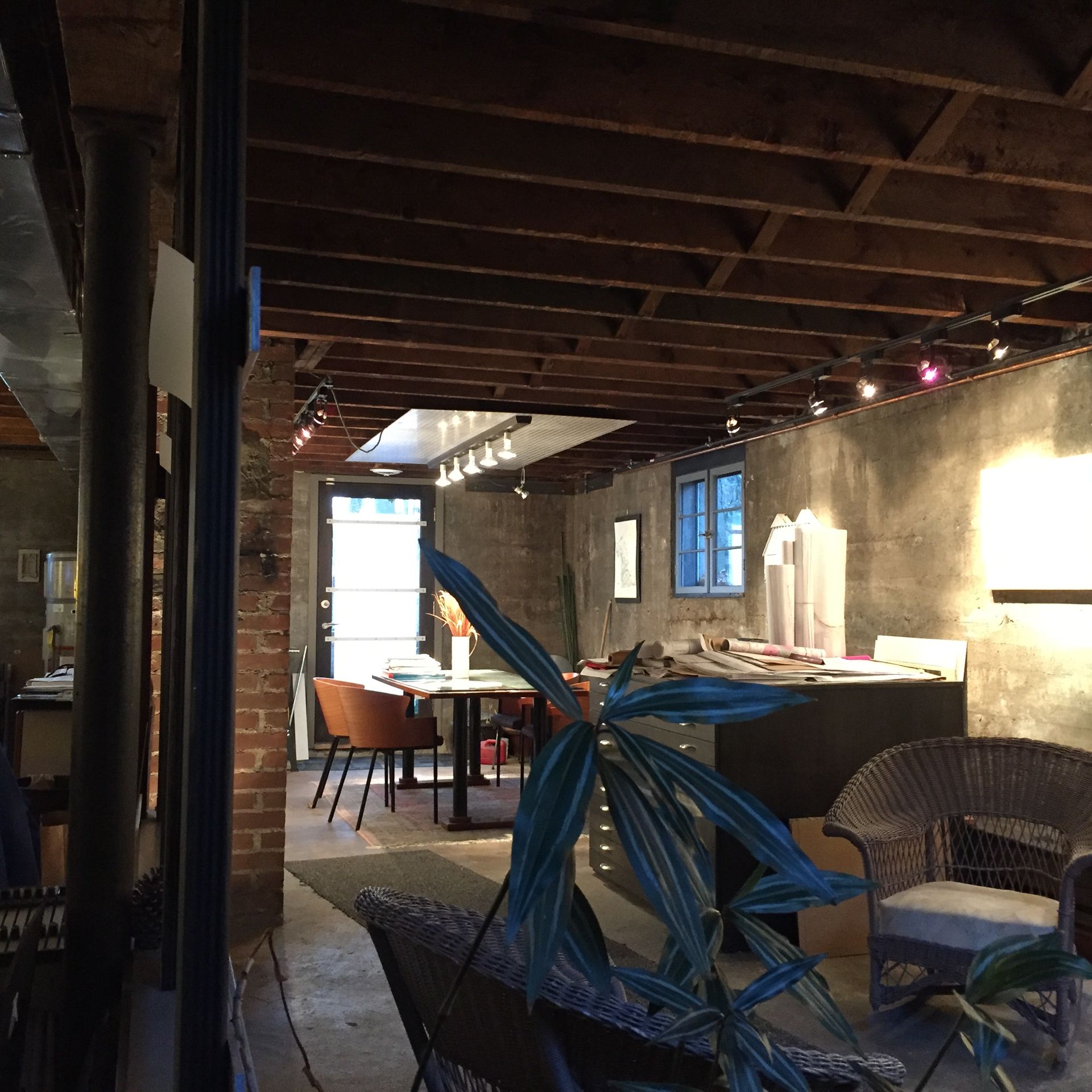 Basement office with exposed beams, brick column, and a round table. Plants and wicker chairs are in foreground.