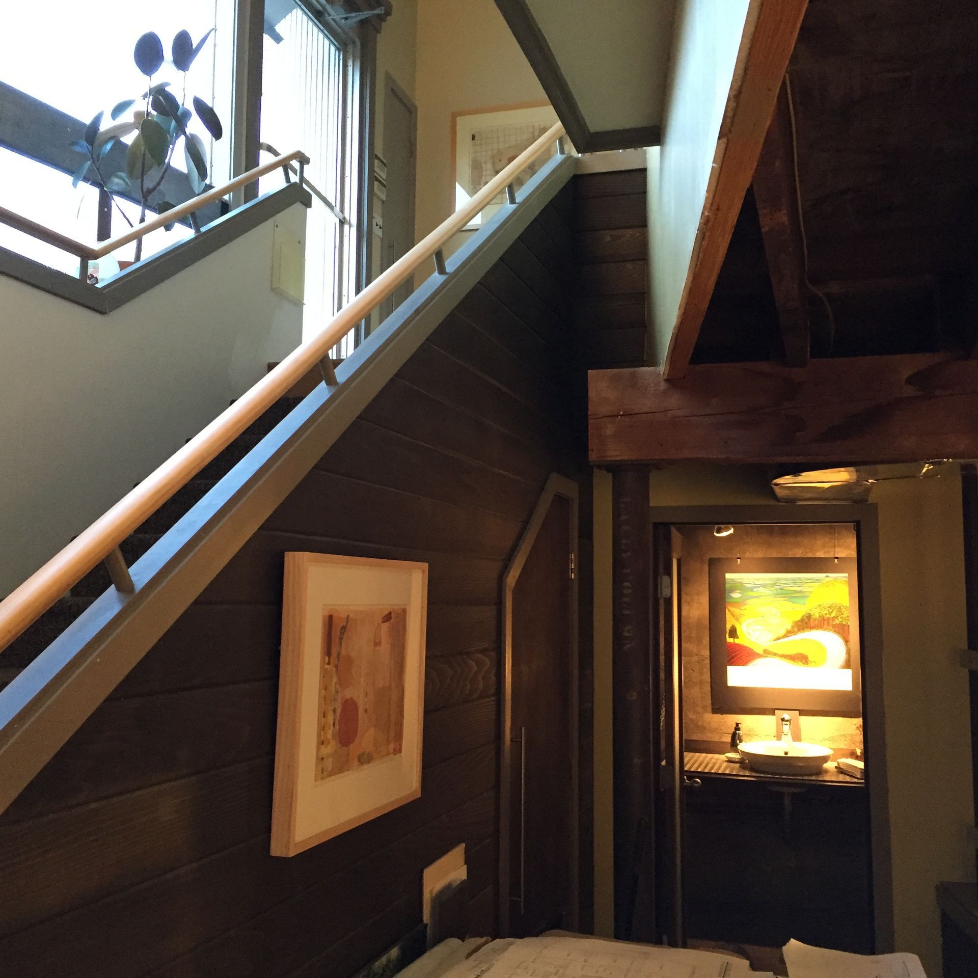 Staircase with wooden paneling, a handrail, and a small painting; an open doorway to a room with a sink and art.