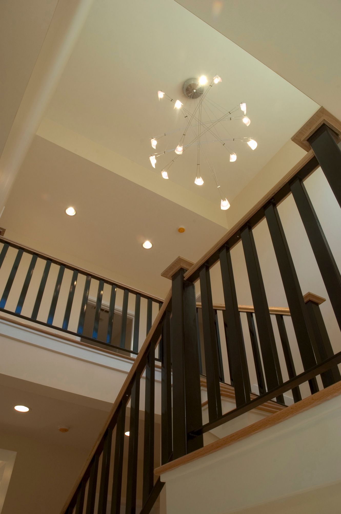 Staircase with black railing and light-colored wooden accents, leading up. Overhead, a cluster of hanging lights.