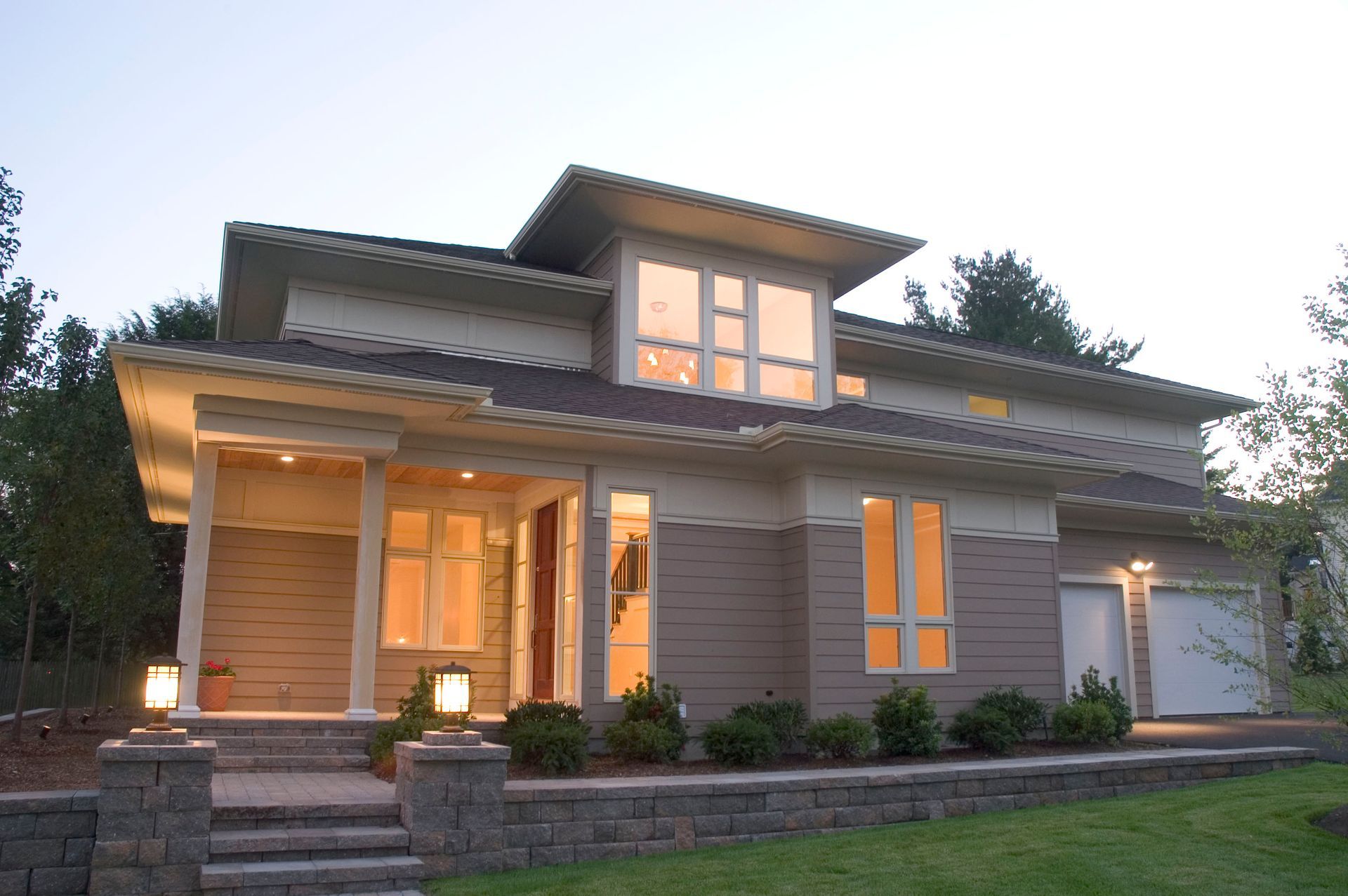 Modern two-story house with tan siding, large windows, and a covered porch; illuminated at dusk.