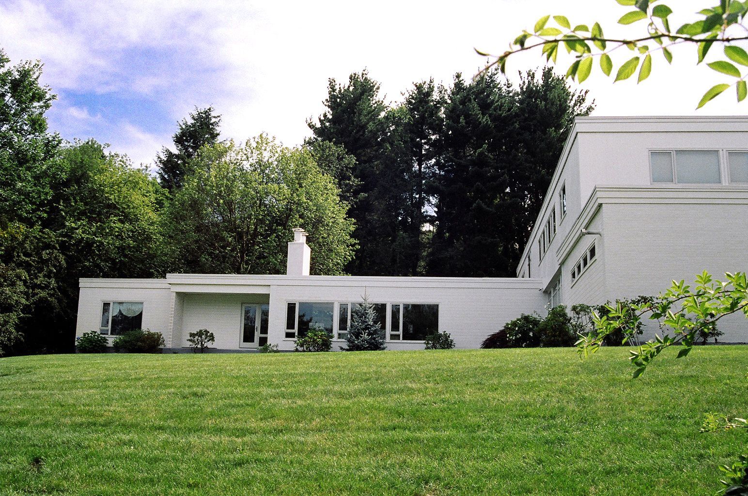 White modern house with flat roof, large windows, and green lawn. Trees in background.