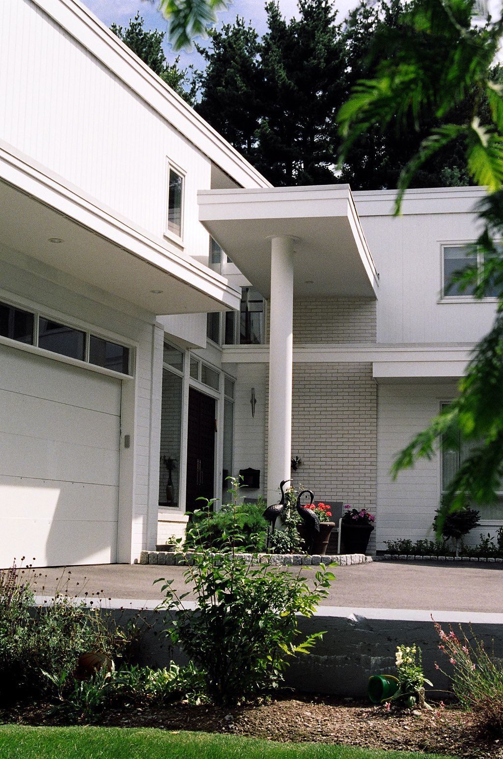 White modern house with a carport and a columned entrance, plants in the foreground, and a paved driveway.
