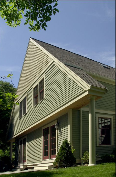 Green-sided house with a steeply angled roof, windows, and a porch, set in a yard with a blue sky.