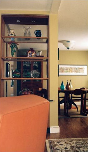 Orange sofa next to wooden bookshelf displaying glassware. Dining room with a table and artwork visible.