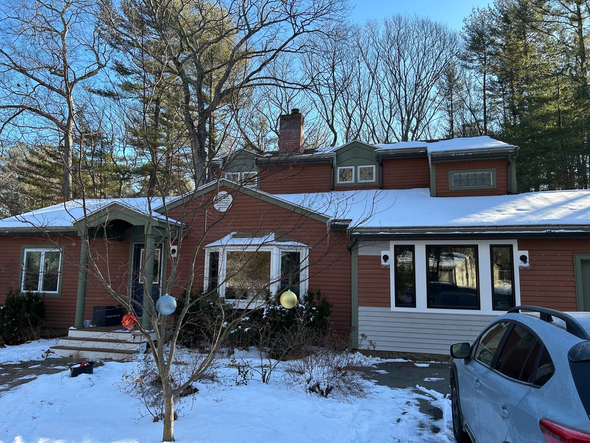 Brick house with snow-covered roof and yard; parked car in the driveway, trees in the background.