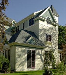 Light yellow house with teal and green trim, with a tower featuring a round window, under a clear blue sky.