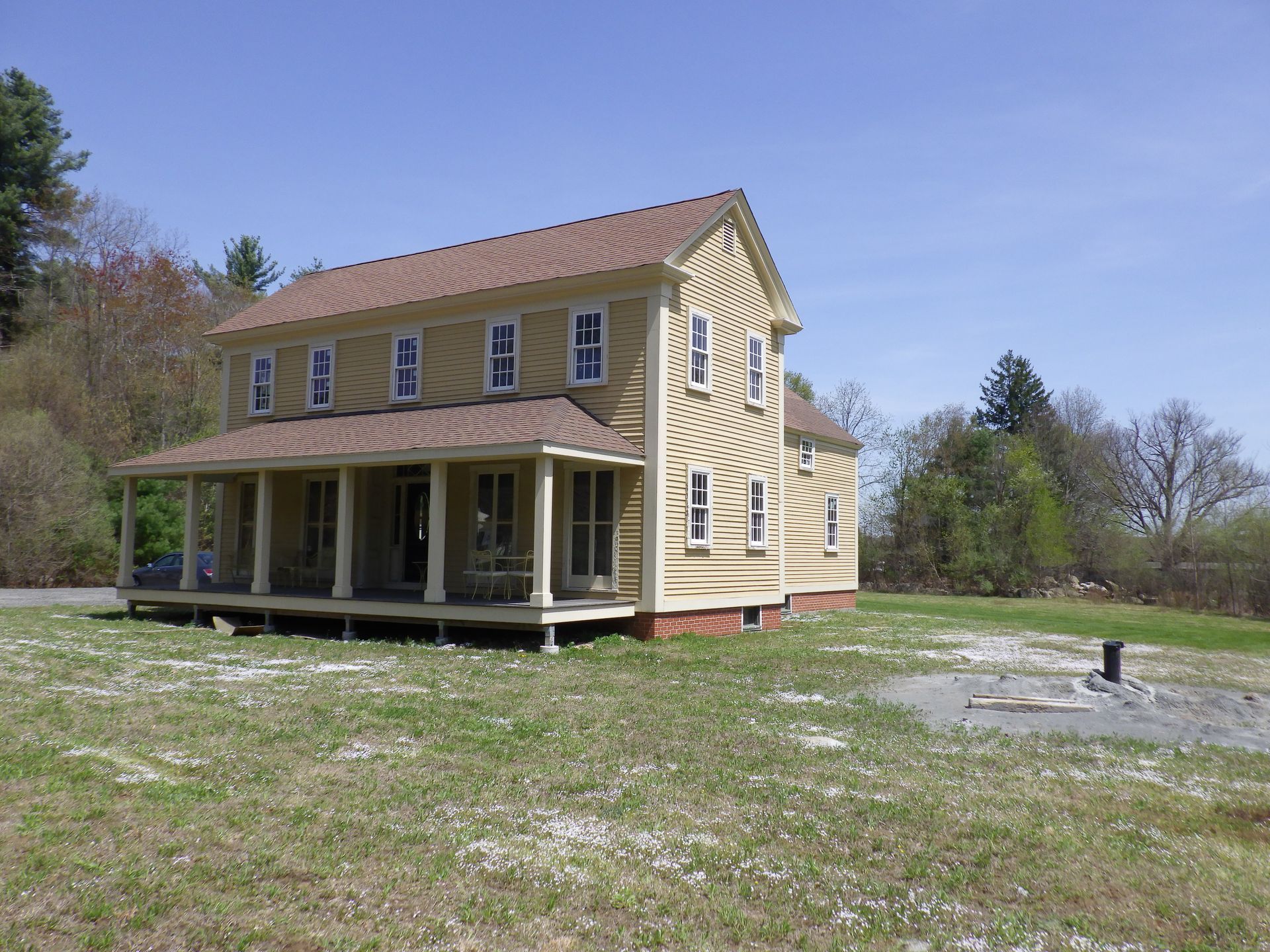 Two-story, light yellow house with porch. Brown roof and trim. Situated on grassy lot with trees and blue sky.