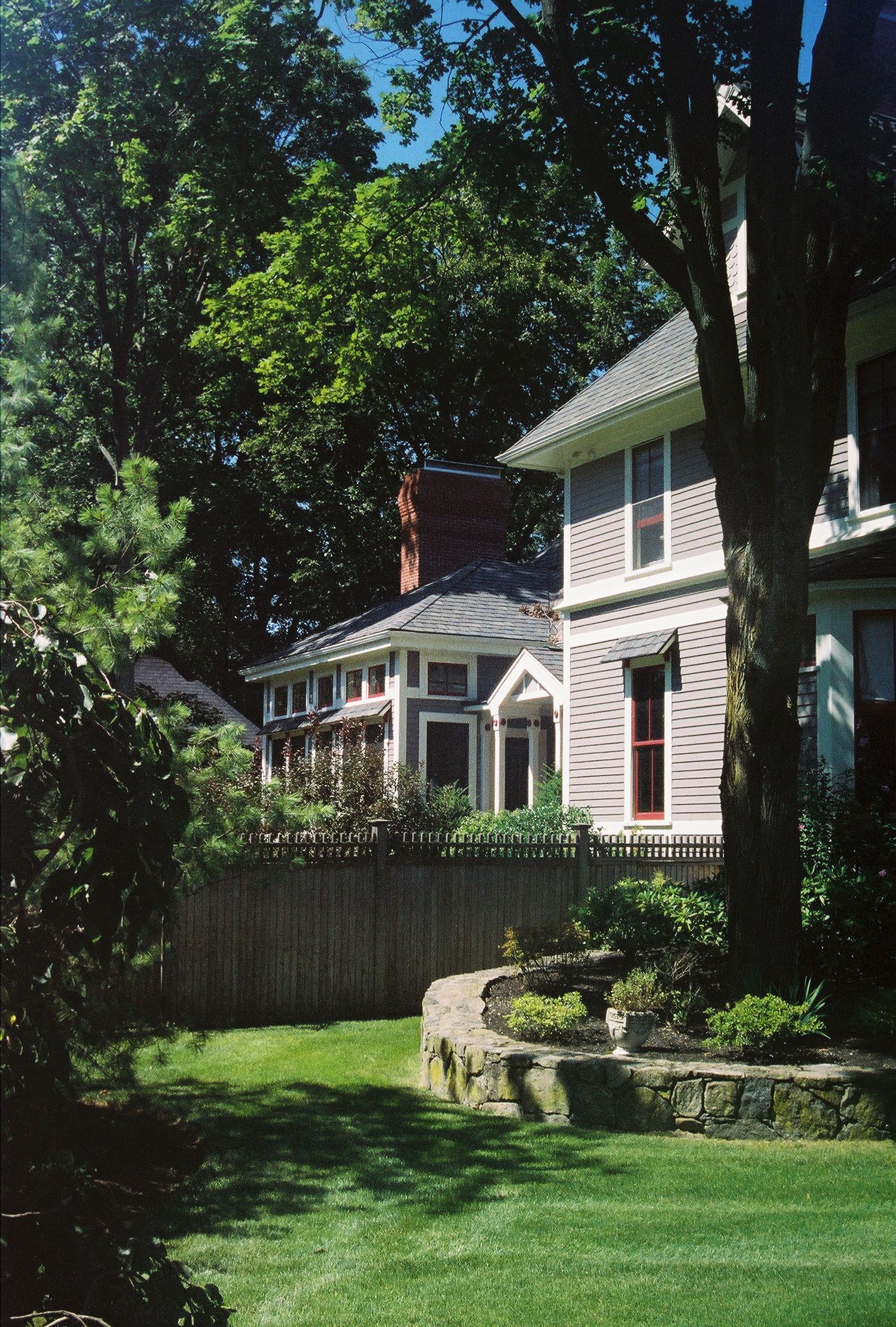 Houses behind a wooden fence, seen from a green yard with a tree and stone landscaping.