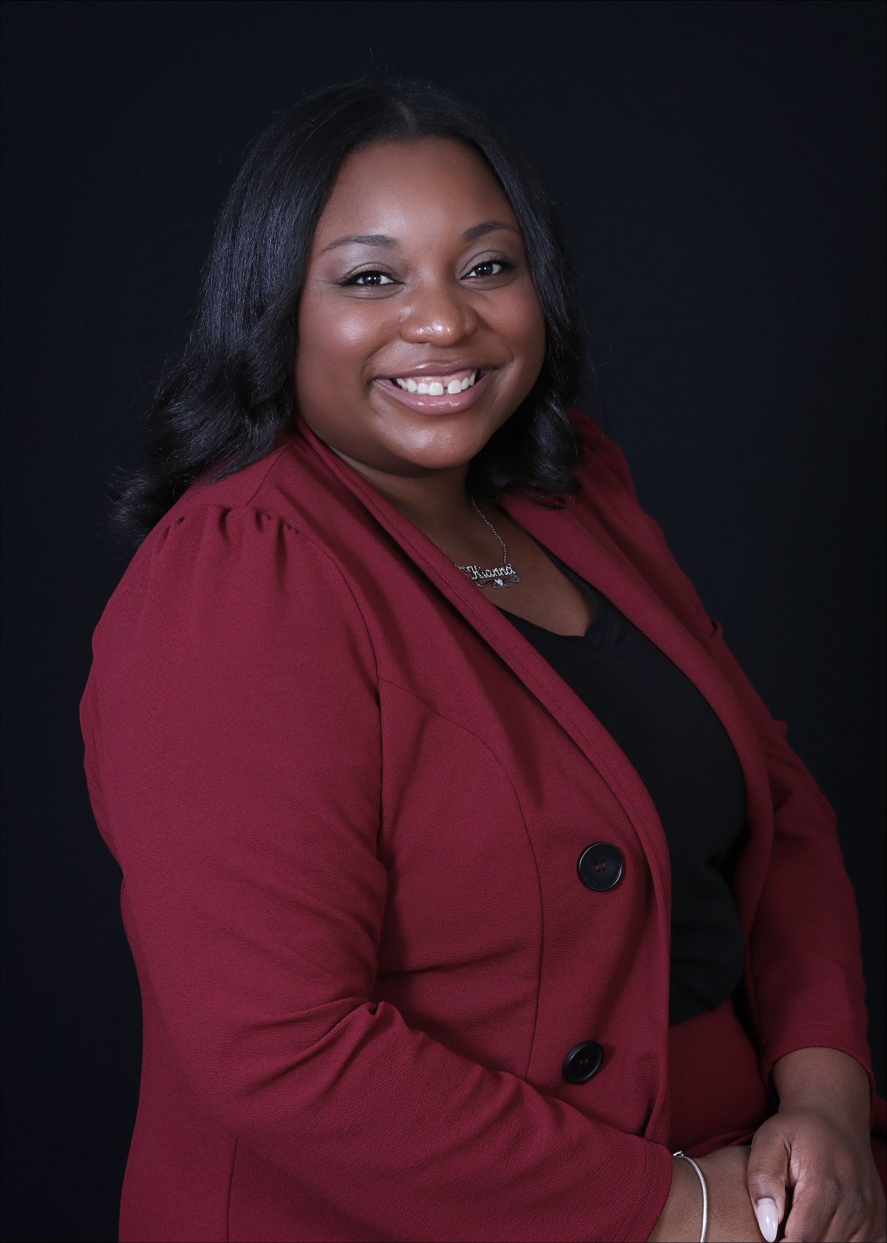 Kianna White with dark hair, smiling, in a maroon blazer and black top, against a black background.