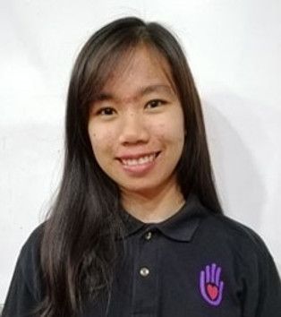 Young person smiles, wearing a black polo shirt with a purple hand logo, against a white wall.