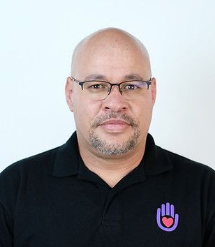 Bald man wearing glasses, black shirt with purple heart/hand logo. Neutral expression.
