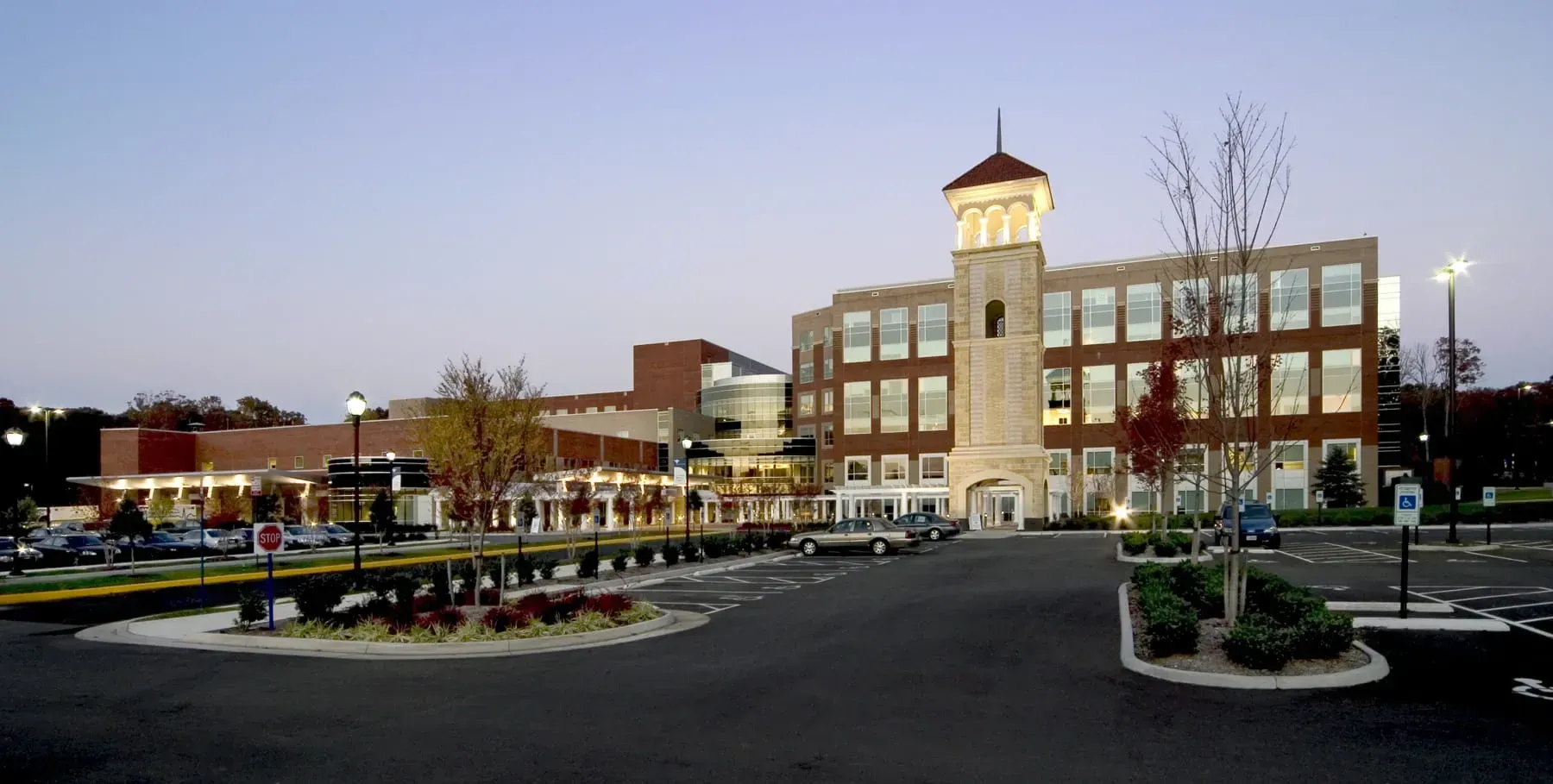 A multistory brick office building with a central clock tower overlooking a parking lot at dusk.