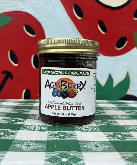 A jar of Agriberry apple butter sitting on a green and white checkered tablecloth in front of a strawberry-patterned wall.